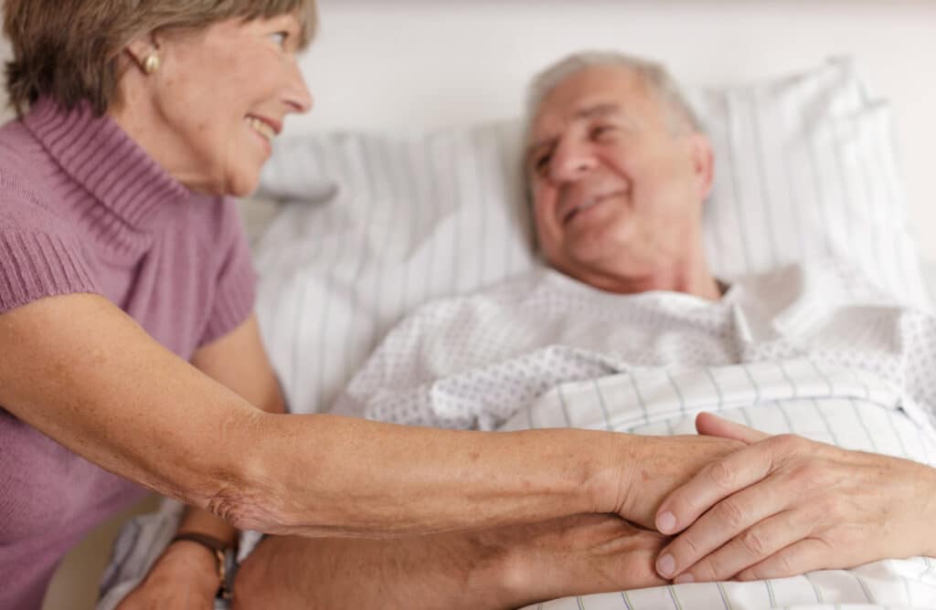 Wife comforting husband laying in hospital bed.