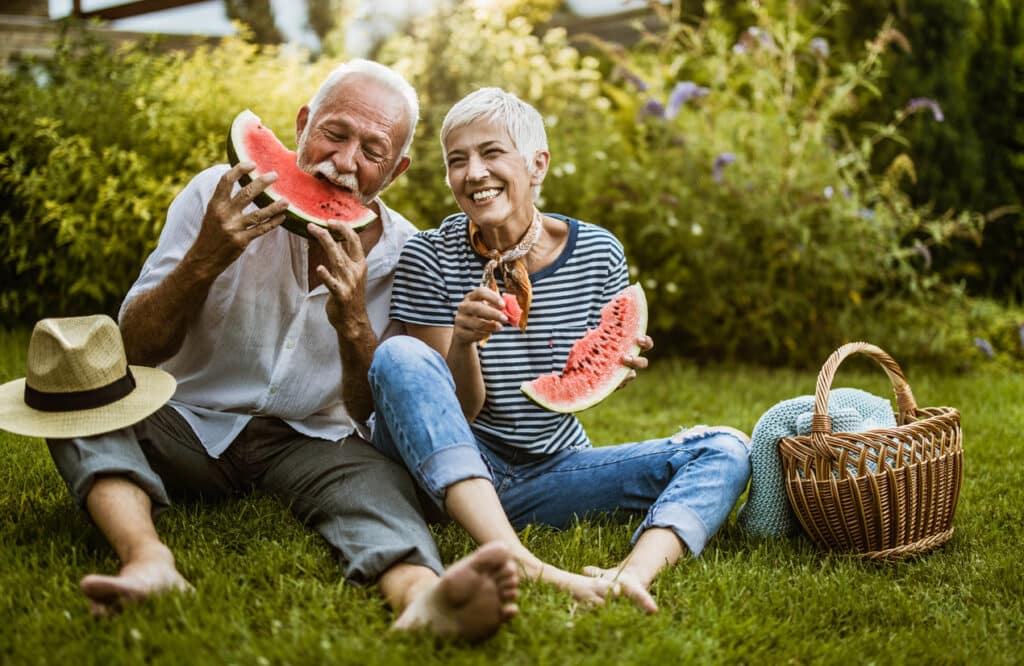 Cheerful mature couple having fun while eating watermelon during picnic day in their backyard.