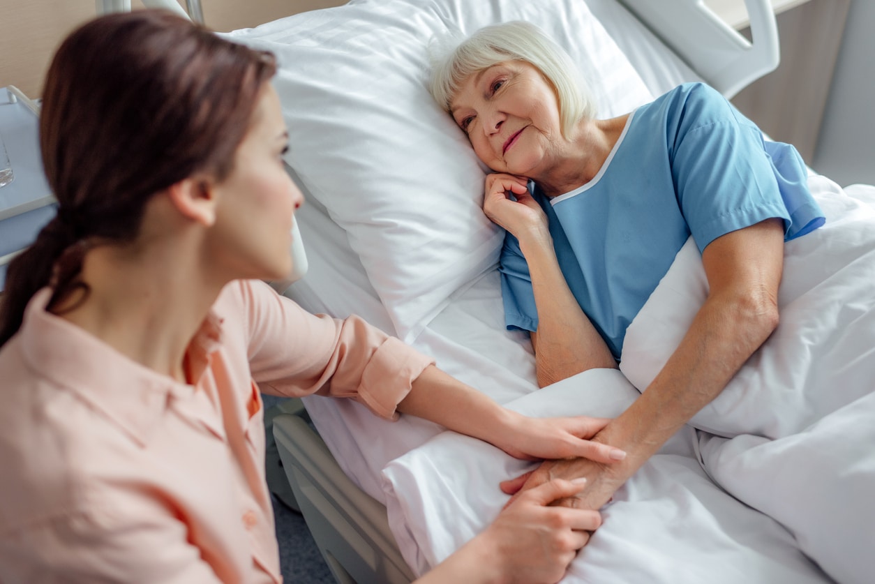 daughter sitting near senior mother in bed and holding hands in hospital - Postoperative Care