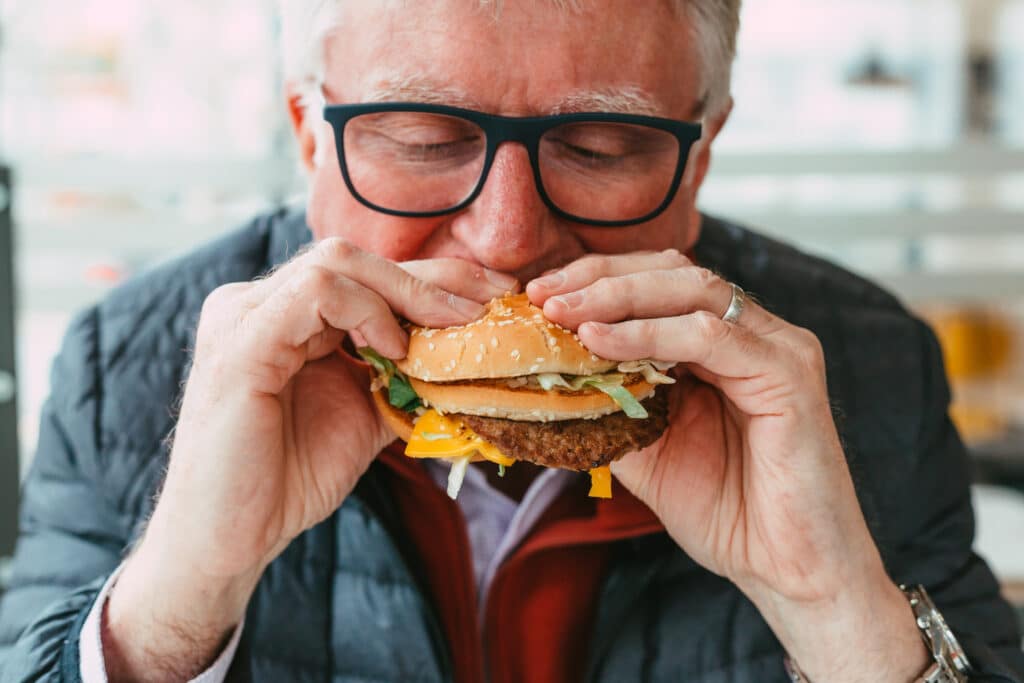 Close up color image depicting a senior man wearing spectacles eating a freshly cooked burger in a fast food restaurant. The burger is messy, with lettuce and cheese hanging out of the bun. Room for copy space.