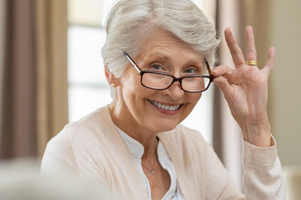 Happy retired senior woman looking at camera while holding eyeglasses. Smiling satisfied woman wearing spectacles at home. Closeup face of old grandmother trying on new eyewear.