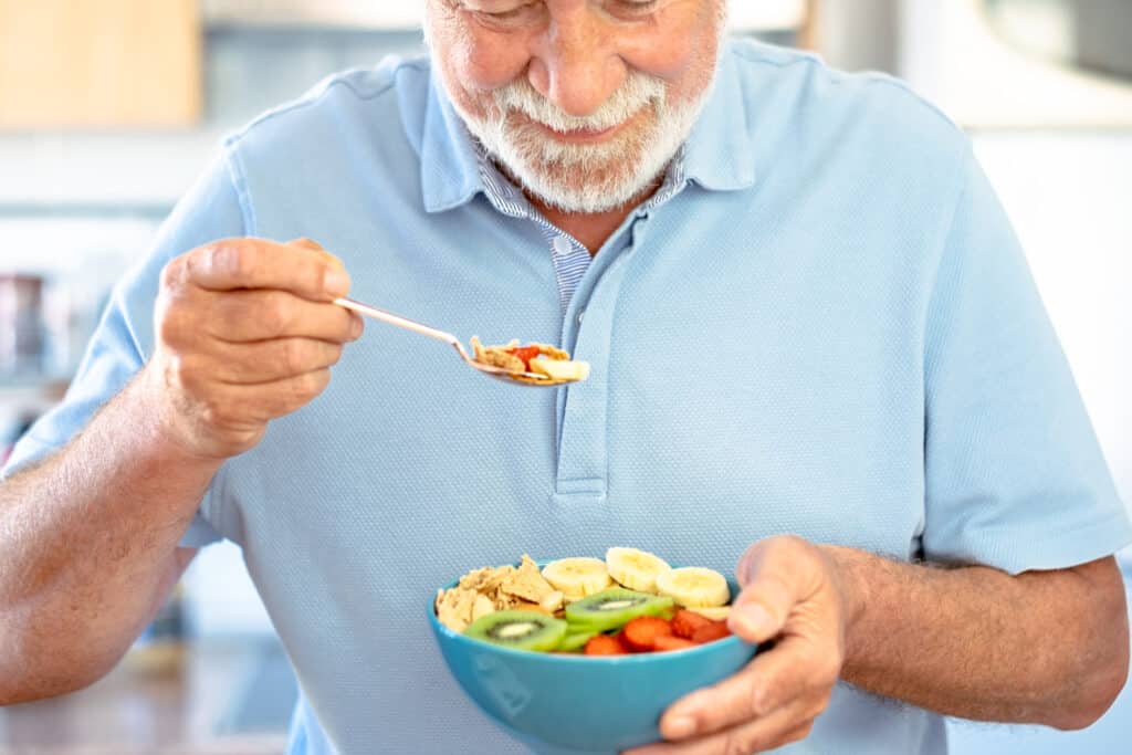 Bearded senior caucasian man holding a ready to eat fruit salad with kiwi banana strawberries and dried fruits. Breakfast or break time, healthy eating
